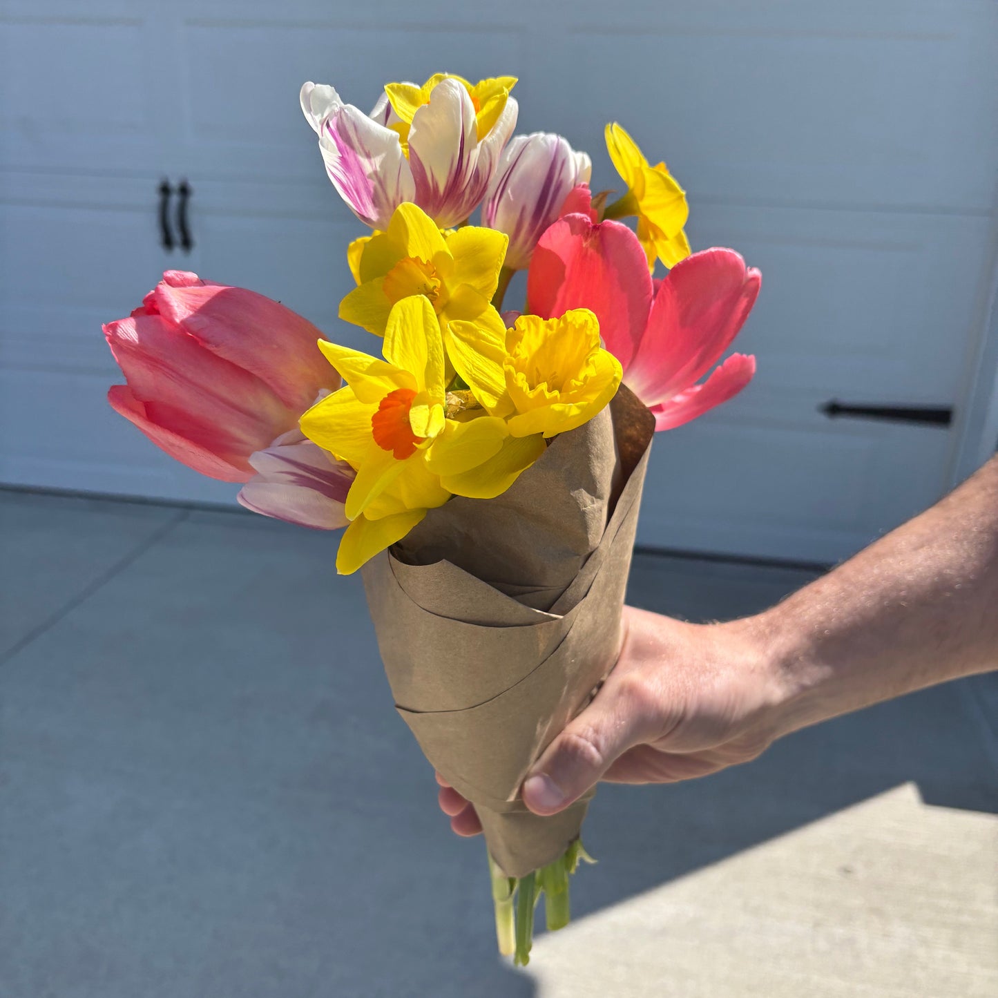 Hand holding a bouquet of flowers wrapped in brown paper against a light-colored wall.