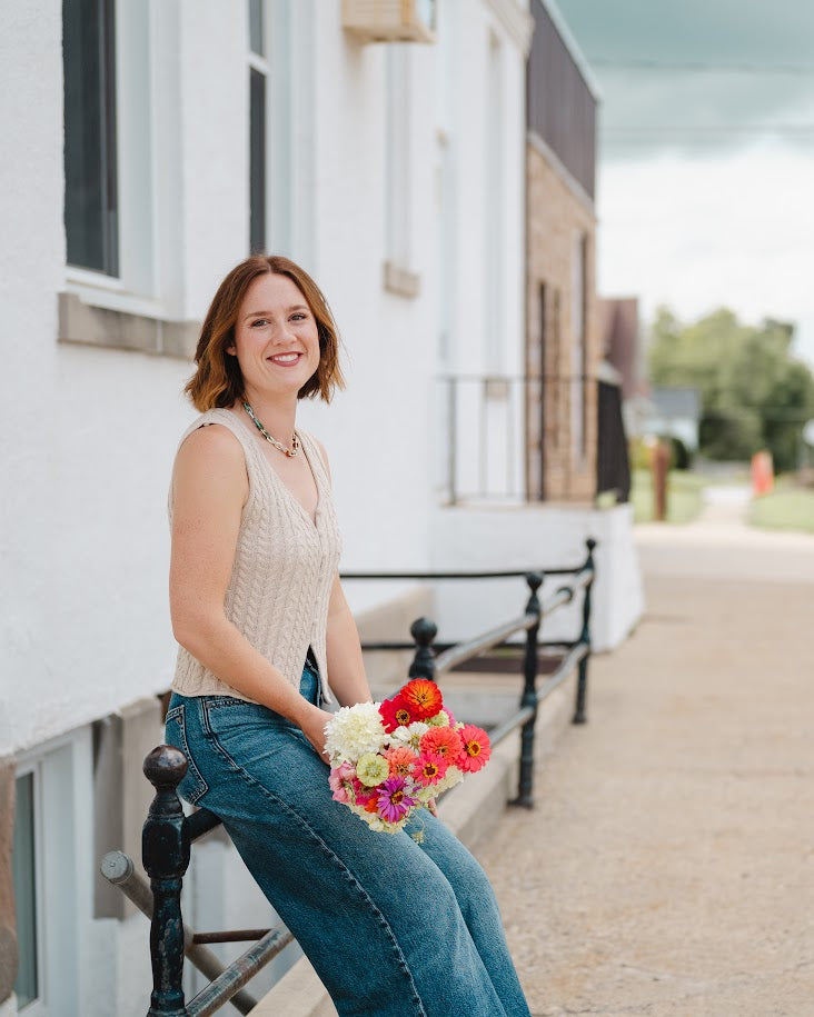 A woman with short brown hair sits on a black metal bench outdoors, smiling and holding a colorful bouquet of flowers. She is wearing a beige sleeveless knit top and blue jeans, with a white building and a gravel path in the background.