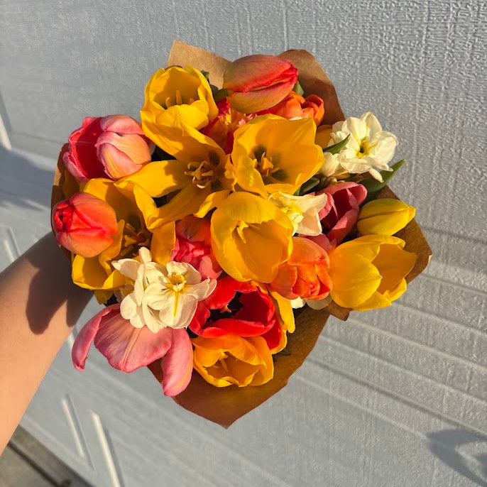 A hand holding a small bouquet of spring flowers, including yellow tulips, pink tulips, and white daffodils, wrapped in brown paper against a white garage door background. Tulips are grown in the Des Moines Iowa area at Rosie Days Flower Farm.