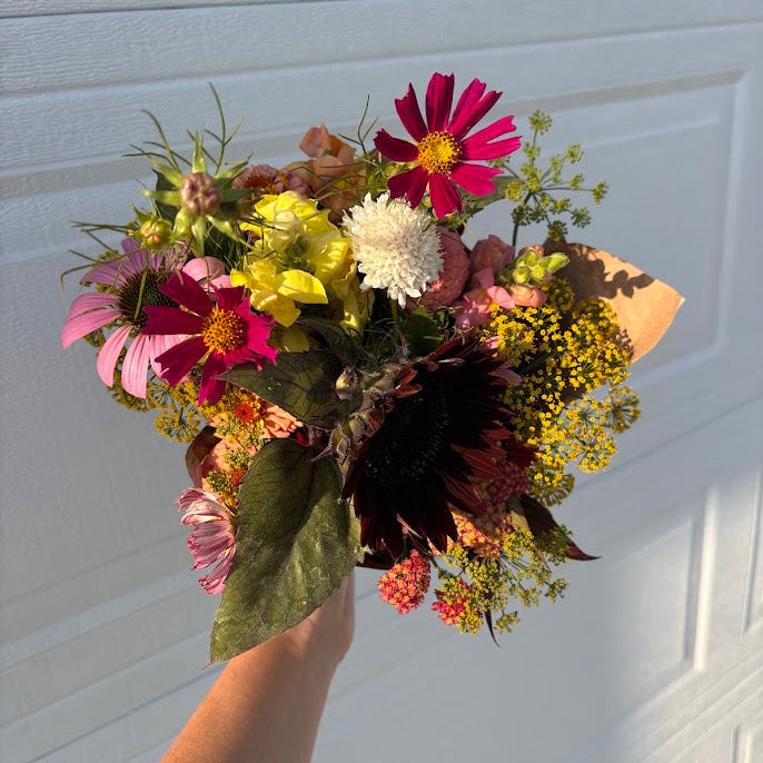A hand holding a rustic, multi-colored wildflower bouquet in front of a white garage door. The bouquet features pink cosmos, yellow flowers, a white daisy, and dark burgundy foliage.