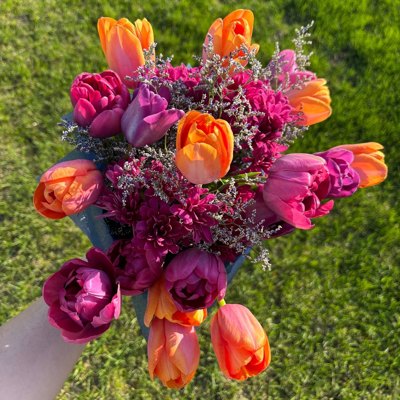 A hand holding a large, vibrant bouquet of mixed spring flowers, including tulips and daffodils, with a green grass background. Flowers locally grown and sold in Ankeny, Iowa.