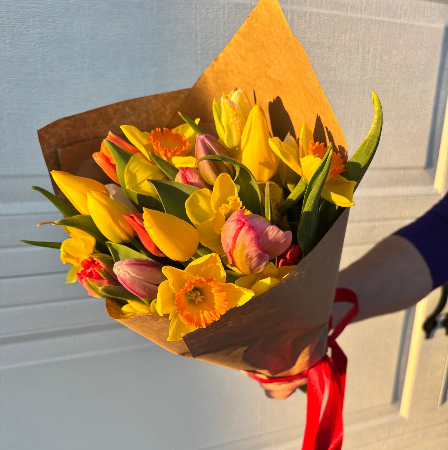 Bouquet of yellow and pink flowers with a red ribbon held against a white door. Bouquet is filled with tulips and daffodils grown at Rosie Days Flower Farm in Ankeny, Iowa. 