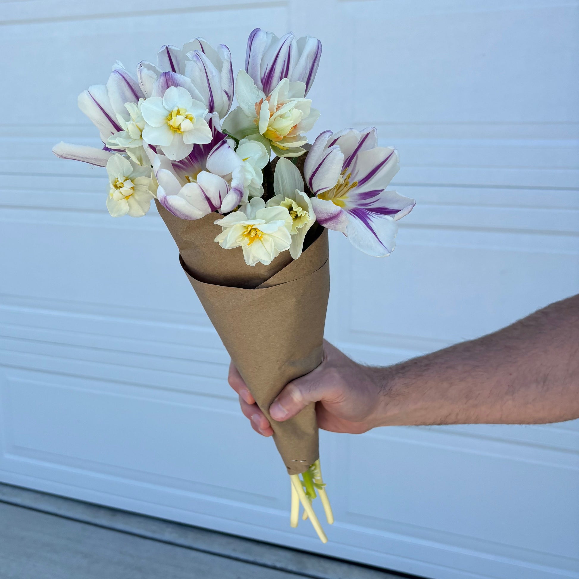 Bouquet of flowers wrapped in brown paper held by a person against a white door. Flowers grown locally at Rosie Days Flower Farm in Ankeny, IA. 