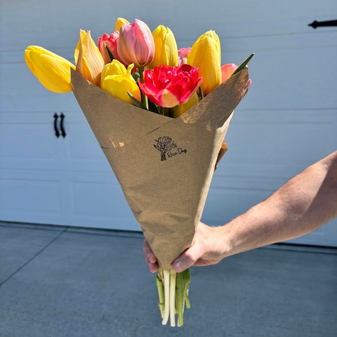 Bouquet of yellow and pink tulips in brown paper wrap held by a person in front of a garage door. Tulips are grown locally at Rosie Days Flower Farm in Ankeny, Iowa.